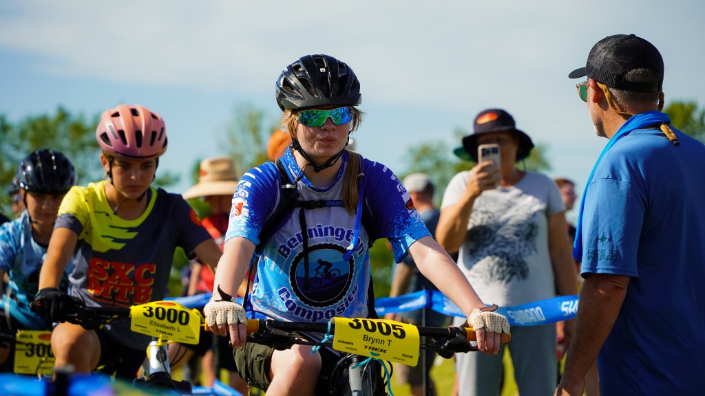 A group of young mountain bikers preparing for a race, with a focus on a girl in blue cycling attire and sunglasses, holding her bike. Surrounding participants and spectators are seen in the background.