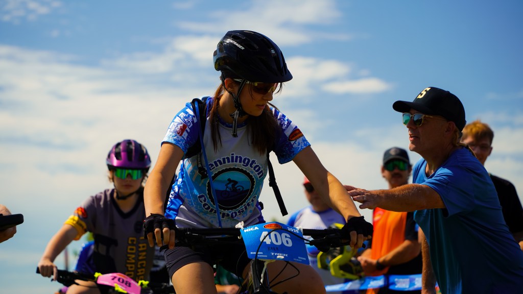 A young female cyclist wearing a helmet and race jersey is preparing for a race while receiving last-minute advice from an adult mentor. In the background, other cyclists are getting ready, set against a clear blue sky.