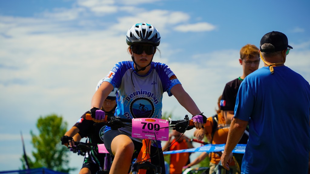 A young female cyclist wearing a helmet and sunglasses prepares to race on her mountain bike, with a number plate attached to her bike. Various people are around her, including race officials and other participants, against a backdrop of trees and a blue sky.