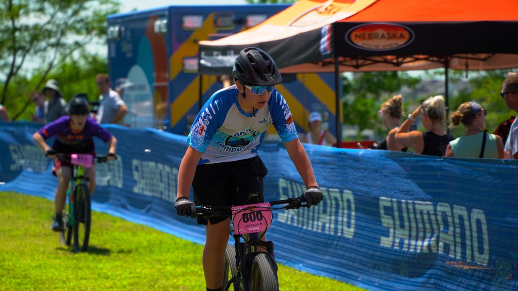 A young cyclist in a blue and white jersey races in a mountain biking event, with a pink number bib on their front. Other participants and spectators are visible in the background.