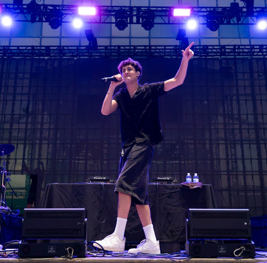 A performer on stage holding a microphone and gesturing with one hand during a music festival, wearing a black shirt and shorts, with speakers and instruments visible in the background.