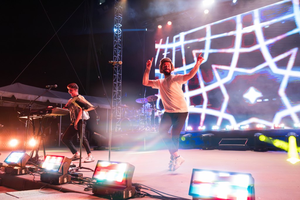 A lively concert scene with two male performers on stage. One is playing a keyboard while the other is dancing enthusiastically, surrounded by colorful stage lights and a vibrant backdrop.