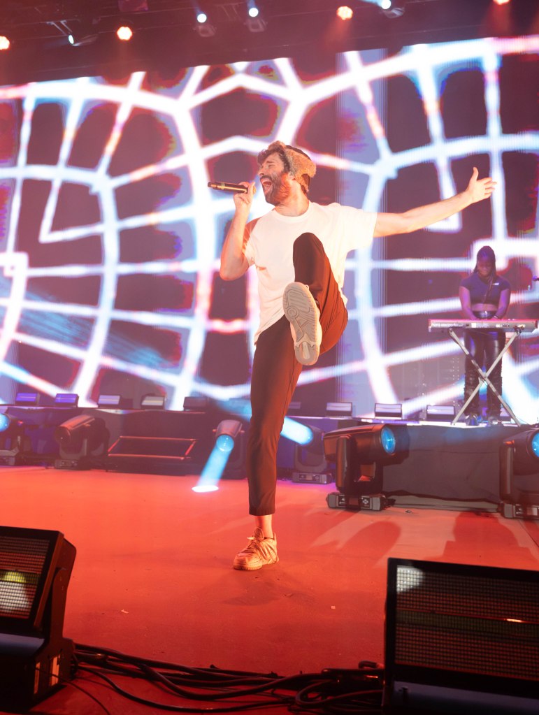 A performer singing on stage with one leg raised, wearing a white t-shirt and black pants, in front of a vibrant light display.