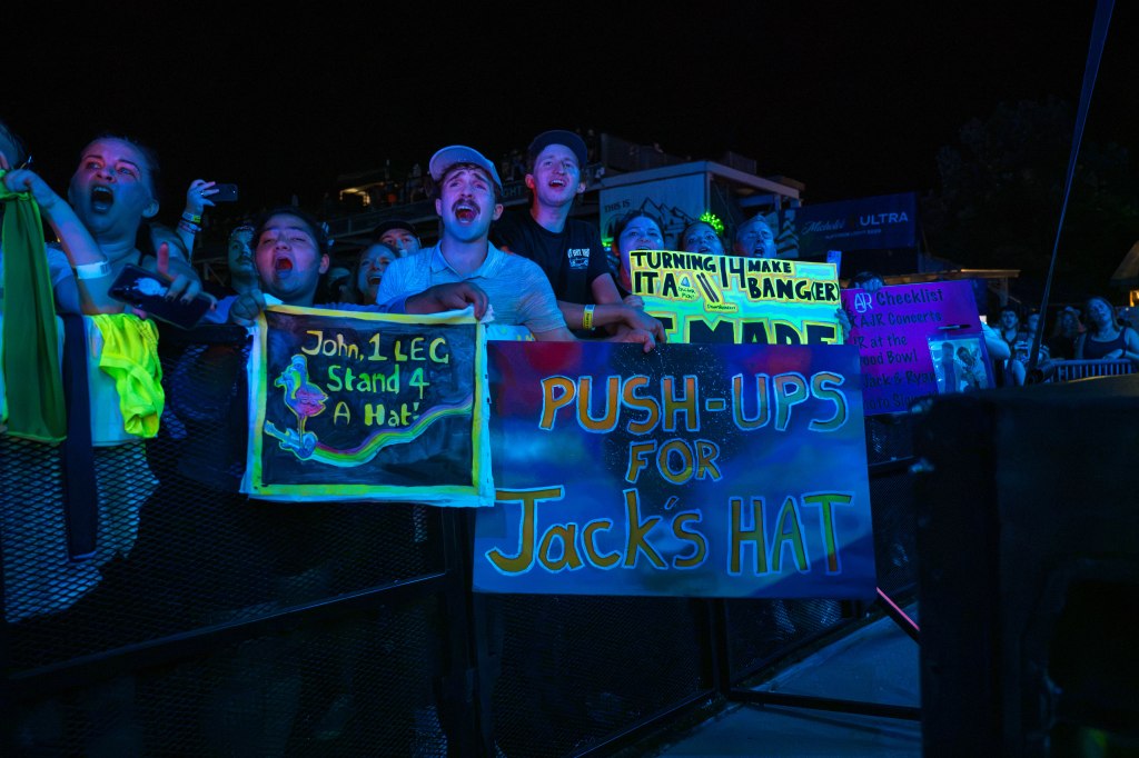 A lively concert crowd cheering with colorful signs, including one that reads 'Push-Ups for Jack's Hat' and another saying 'John, 1 LEC Stand 4 A Hat.'