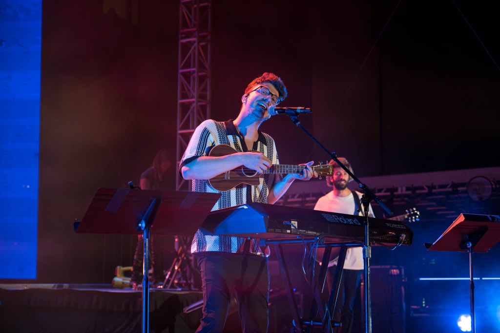 A musician performing on stage with a ukulele and keyboard, surrounded by colorful stage lights and another band member in the background.