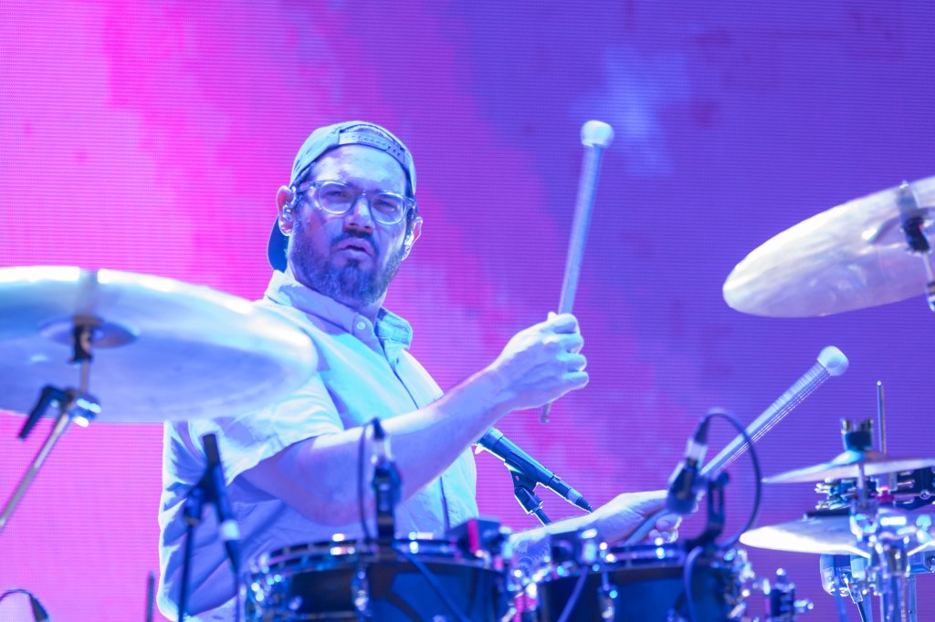 A male drummer performing on stage, wearing glasses and a cap, holding drumsticks in front of a vibrant pink background.