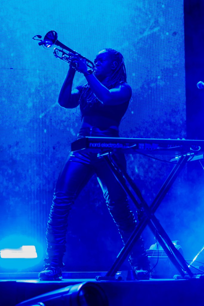 A musician performs on stage, playing a trumpet while standing beside a keyboard, surrounded by blue lighting and visual effects.