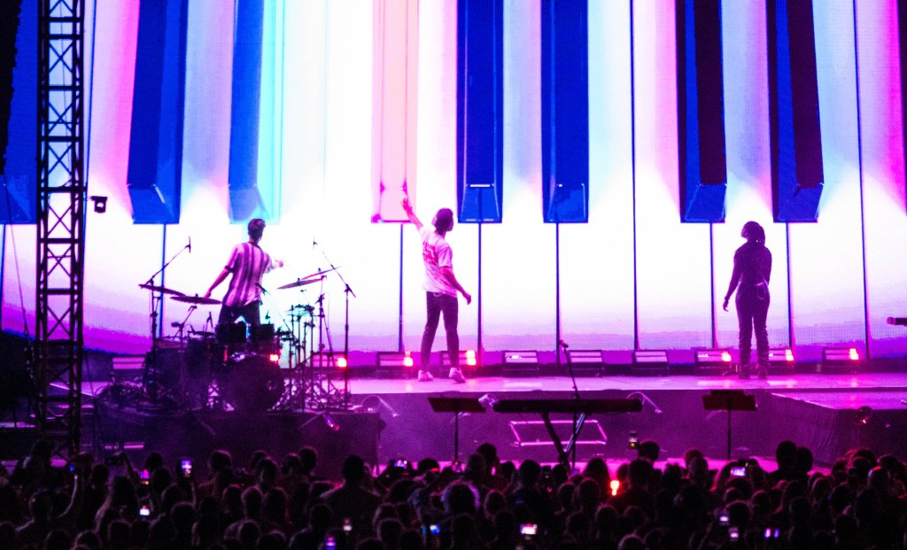 A band performing on stage with a colorful backdrop of piano keys, illuminated by vibrant lights, as an audience watches with their phones raised.