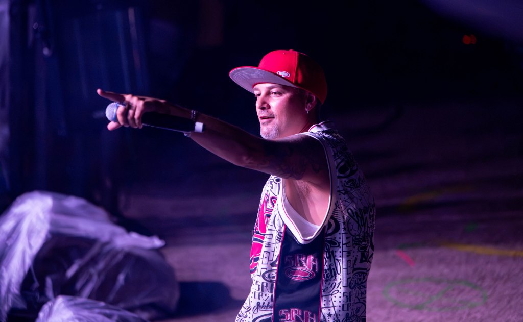 A male performer in a red cap and a sleeveless shirt holds a microphone and gestures while performing on stage.