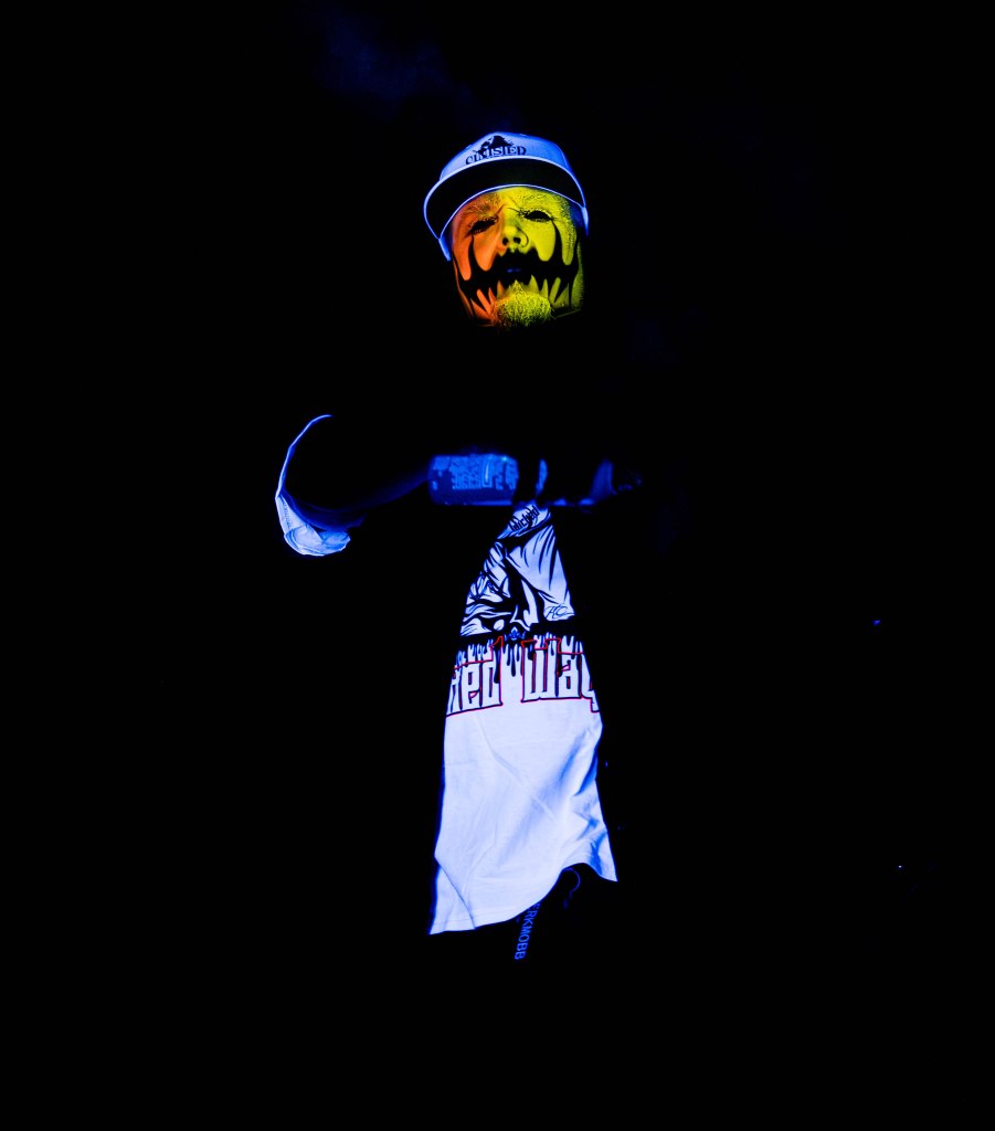 A performer with yellow and black face paint in a dark environment, wearing a cap and a graphic t-shirt.