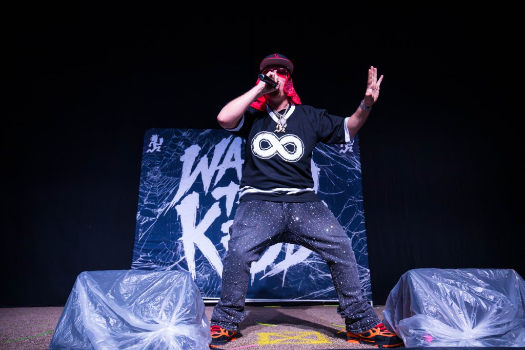 A male performer on stage wearing a black shirt with an infinity symbol, gray pants with a sparkly design, and orange sneakers. He is holding a microphone and wearing a red bandana while performing.