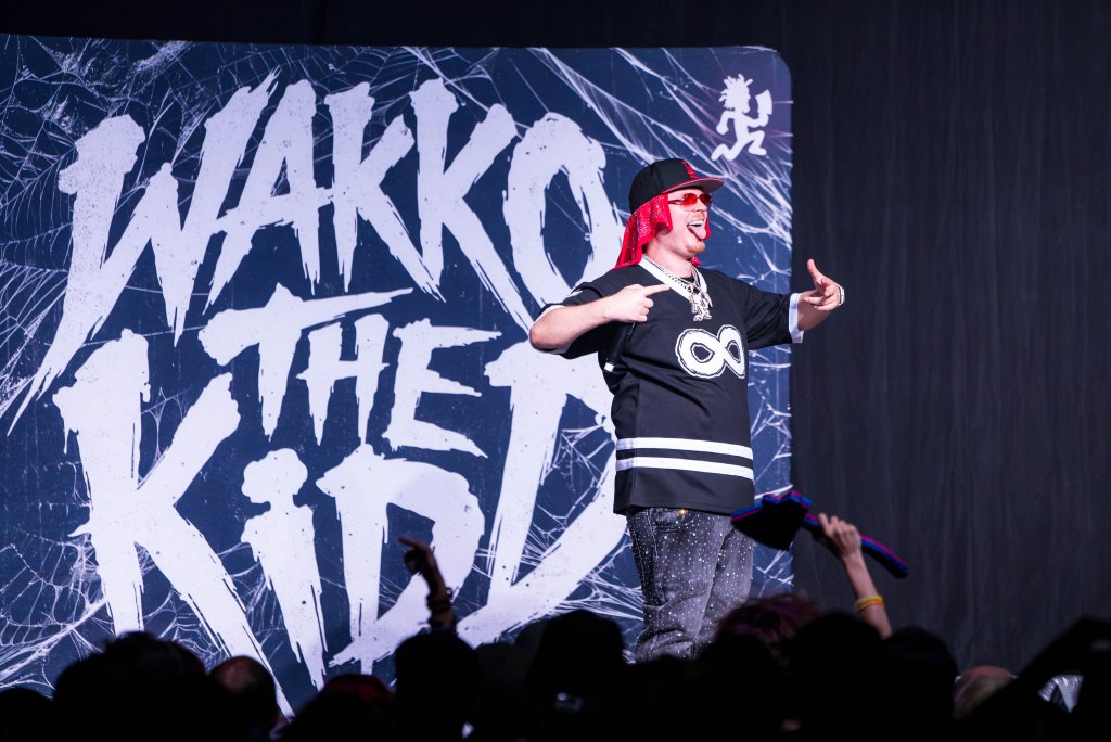 A performer with red hair and sunglasses strikes a playful pose on stage in front of a large backdrop displaying the name 'Wakko the Kidd'.