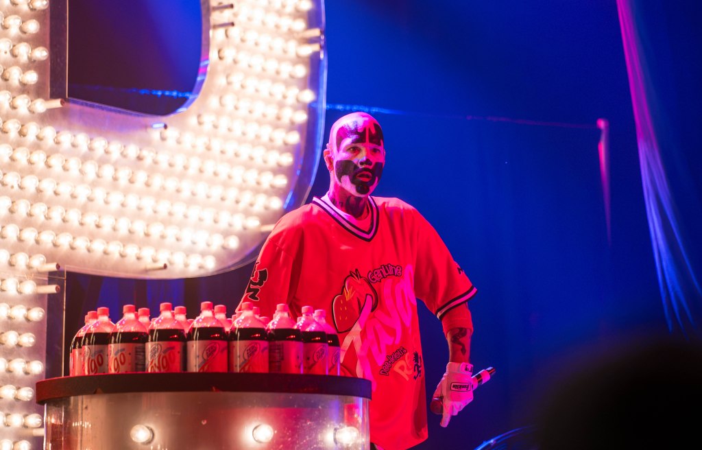 A performer with face paint wearing a red jersey stands in front of a bright display with bottles of soda, illuminated by colorful stage lights.