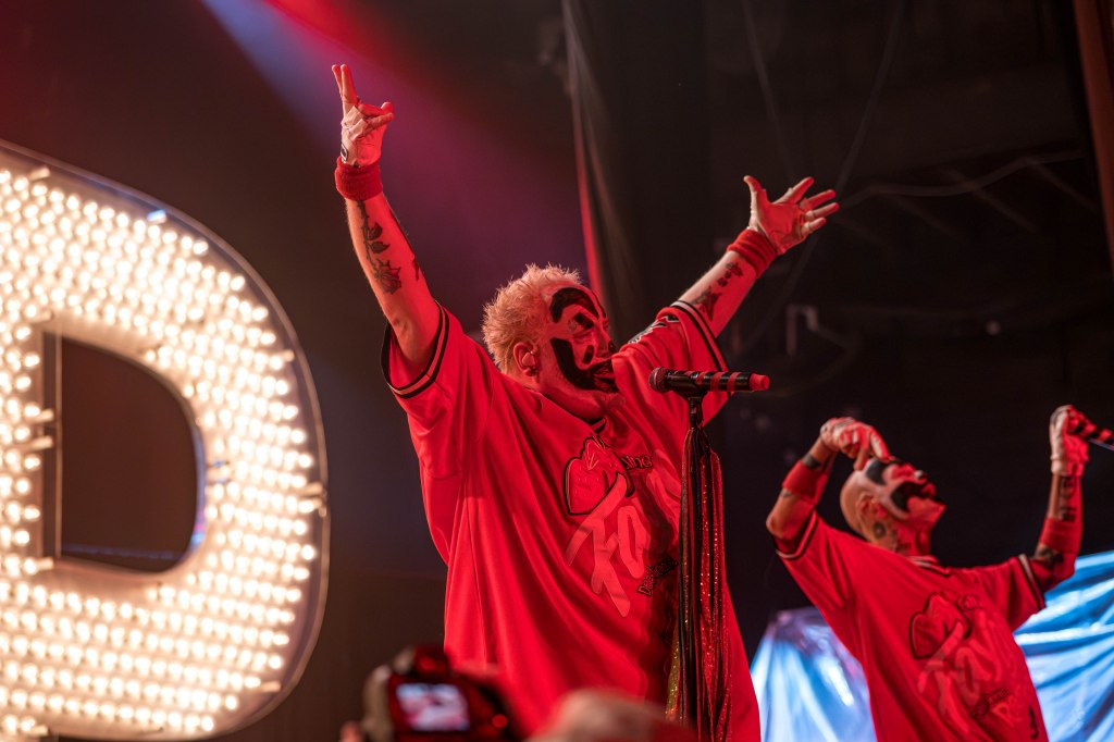 Two performers in clown makeup and matching red jerseys are energetically engaging with the audience on stage, with one raising both arms and the other holding a microphone.