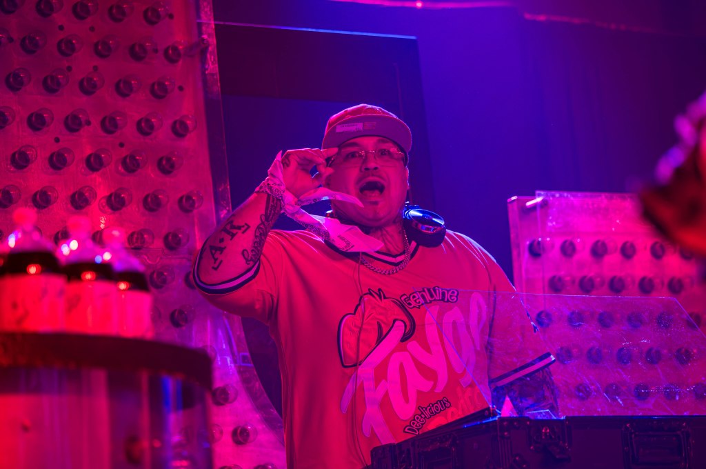 A DJ wearing a colorful shirt with headphones around his neck, gesturing with his hand while performing on stage. The background features vibrant lighting and bottles, creating an energetic atmosphere.