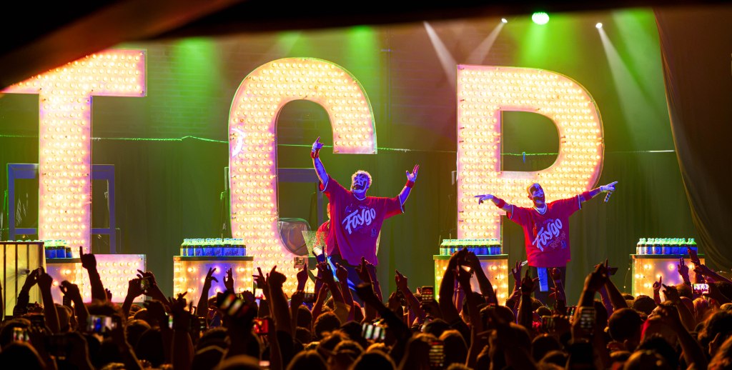 A lively concert scene featuring two performers onstage with large illuminated letters 'TCR' behind them. The crowd in the foreground is enthusiastic, holding up phones and cheering.
