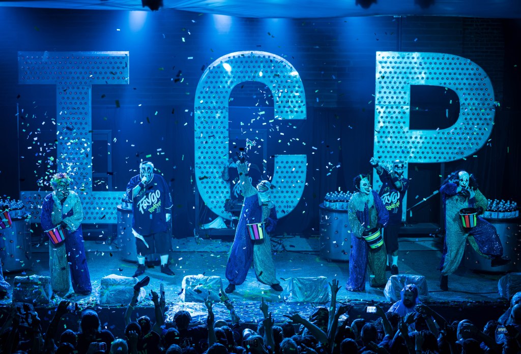 A performance scene featuring five entertainers in colorful costumes and face paint, standing on stage with large, illuminated letters 'TCP' behind them. Confetti is falling from above, while the audience in the foreground appears excited.