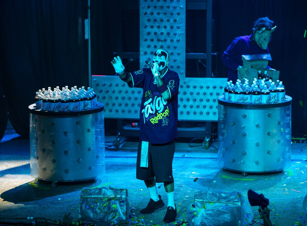 A performer in a colorful outfit stands on stage, gesturing while speaking into a microphone. Behind him are two circular structures stacked with bottles of soda, and another performer in a mask is visible in the background.