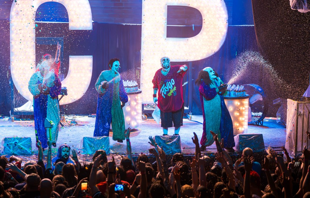A lively performance with four entertainers in clown costumes on stage, covered in colorful paint and confetti, with large illuminated letters in the background. The audience is energetically cheering from the front.