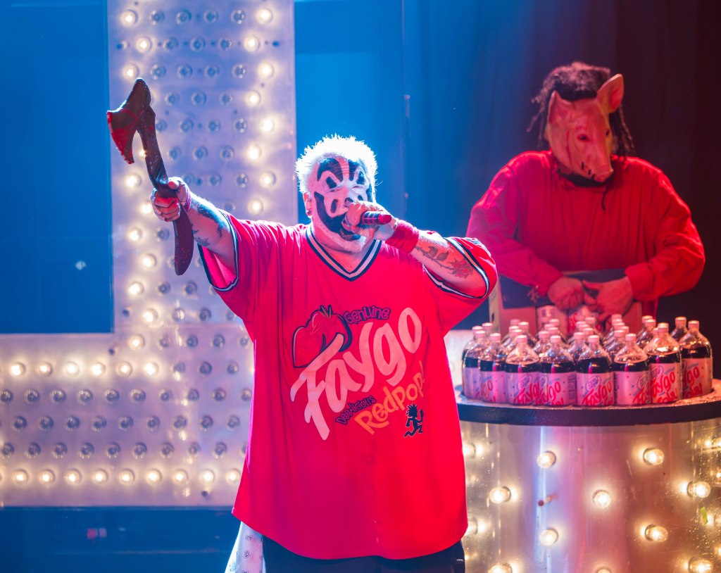 A performer in a red oversized shirt with a Faygo logo holds an axe and sings into a microphone, while another performer in a pig mask stands behind a table with bottles of Faygo soda.