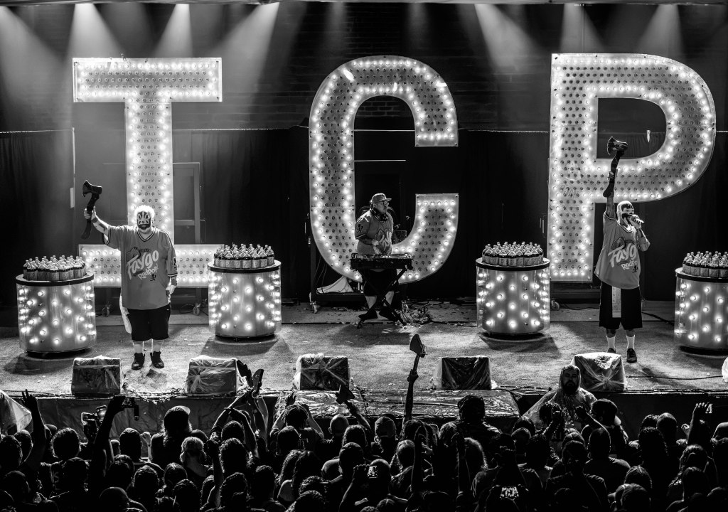 Black and white image of a live concert scene with two performers in front of large illuminated letters 'TCP'. One performer is holding a hat and the other is singing, while a DJ is behind them. The stage is adorned with bottles and the audience is visible in the foreground.