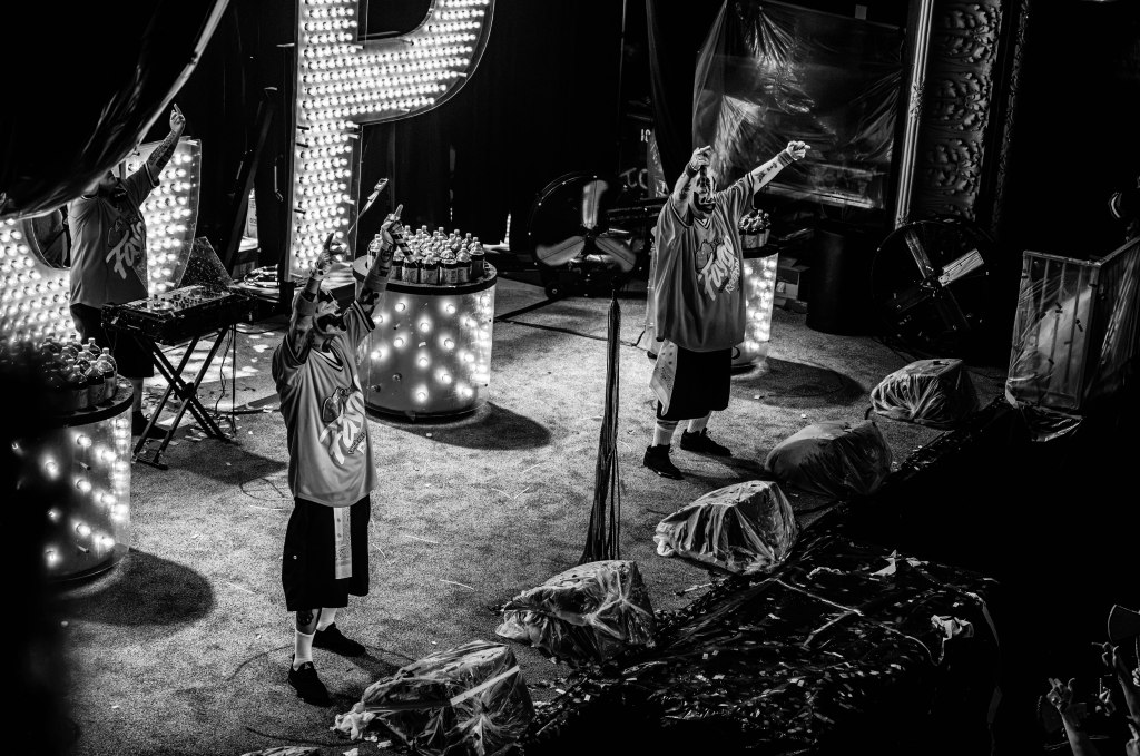 Two performers on stage in black and white, one raising their fist while wearing oversized shirts and shorts, amid illuminated decorative elements and stage equipment.