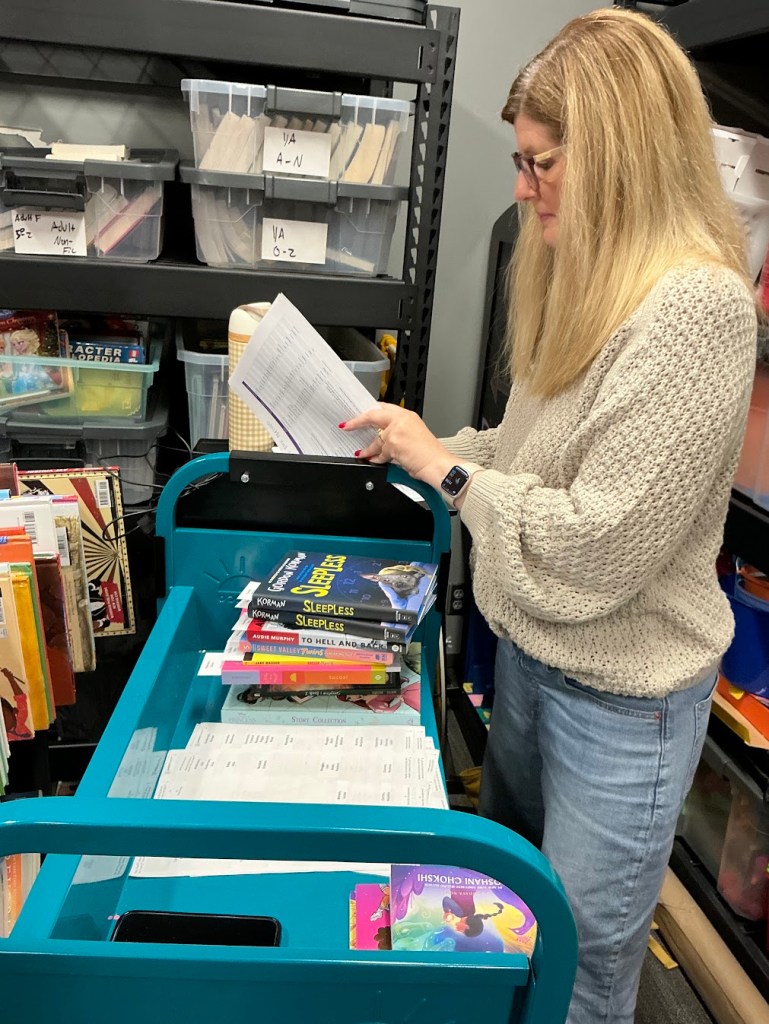 A woman is reviewing papers while standing next to a cart filled with books in a library or storage area, with plastic storage bins visible in the background.