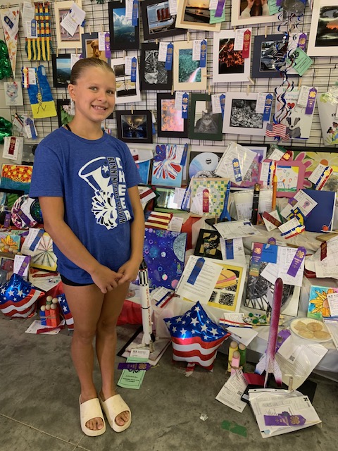 A young girl wearing a blue cheerleading t-shirt poses in front of a display filled with various colorful art projects, ribbons, and photographs.