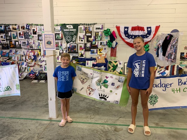 Two young girls standing in front of a display featuring various 4-H projects and exhibits, with colorful posters and decorations. One girl wears a blue cheer t-shirt and stands beside a large banner for the Badger Buddies 4-H Club.
