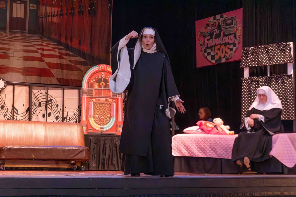 A scene from a stage performance featuring two nuns, one standing and gesturing dramatically while the other sits on a bed. The backdrop depicts a 1950s diner and lockers.