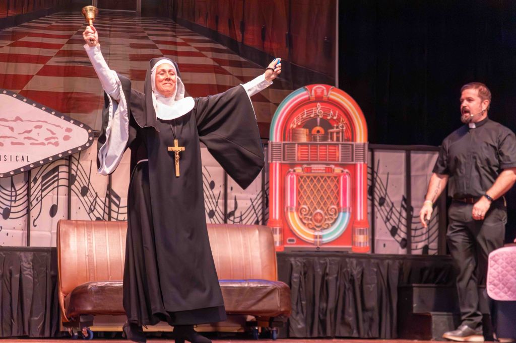 A woman dressed as a nun holds a bell in one hand and raises her other hand in excitement while standing on stage. Next to her is a man in a black clerical shirt, looking at her. The backdrop features musical notes and a vintage jukebox.