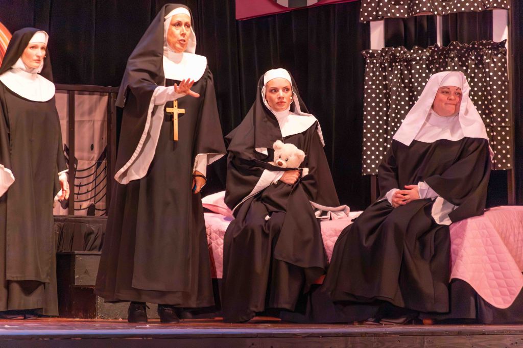 Four nuns in traditional robes performing on stage, with one nun gesturing animatedly, another holding a teddy bear, and two seated. The backdrop features a bed with a pink quilt and polka dot curtains.