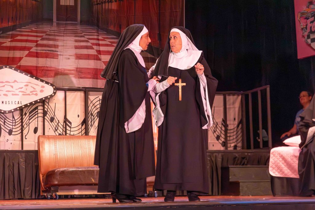 Two nuns engaged in conversation on a stage, with a backdrop of a hallway and musical notes. One nun is holding a cross.