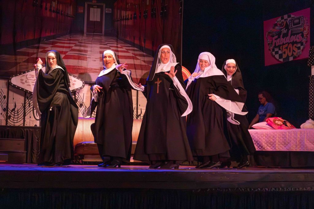 A group of five nuns performing on stage, dressed in traditional habits, with a colorful background featuring musical notes and a 'Fabulous 50s' sign.