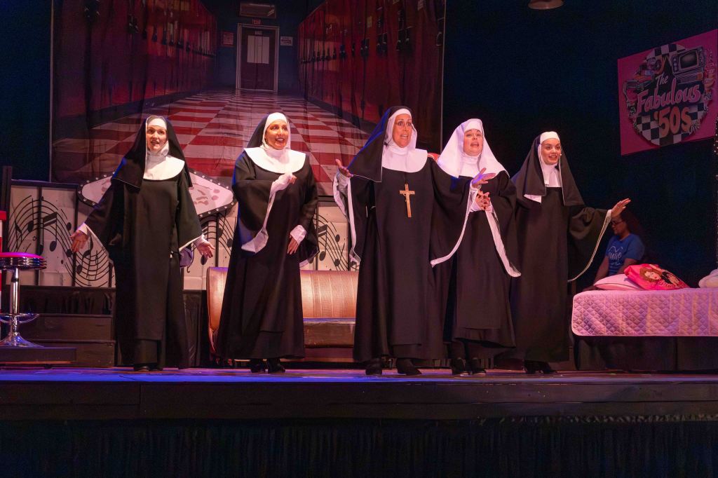 Five nuns in traditional black and white habits performing on stage with a colorful backdrop and musical elements.