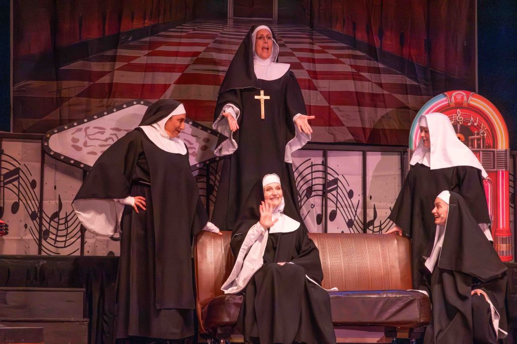 A group of five women dressed as nuns performing on stage, with one nun standing and singing while others sit or interact, set against a backdrop of musical notes and a vintage jukebox.