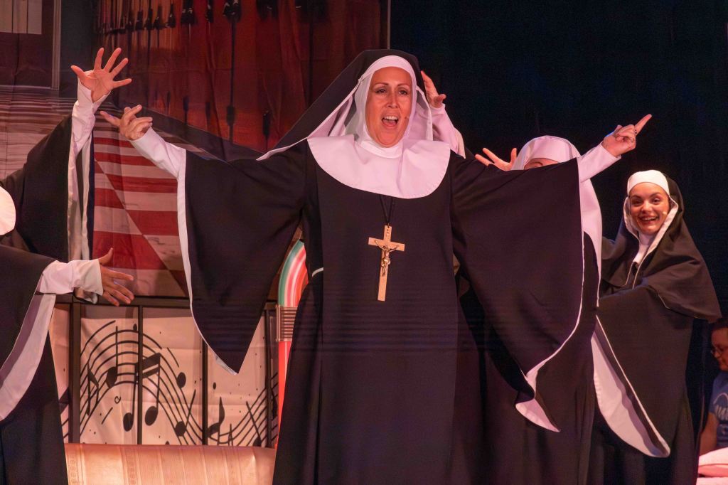 A group of nuns performing on stage, with one nun in the foreground joyfully singing and gesturing, wearing a traditional black habit and a cross necklace.