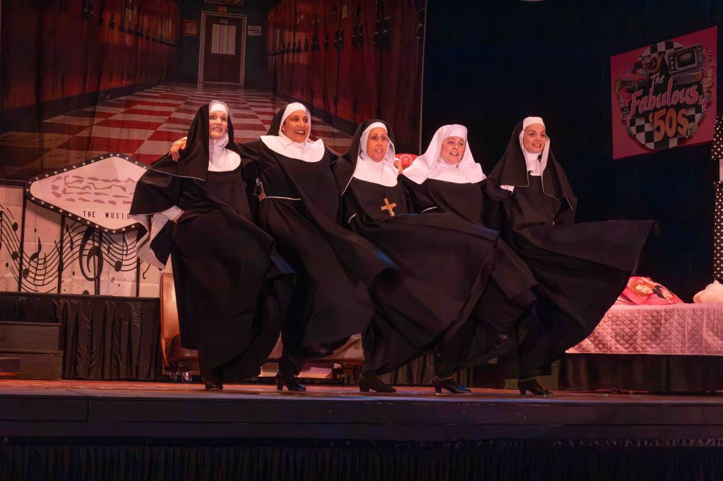 Five nuns in traditional costumes perform a high-kicking dance on stage, with a colorful backdrop depicting a 1950s theme.