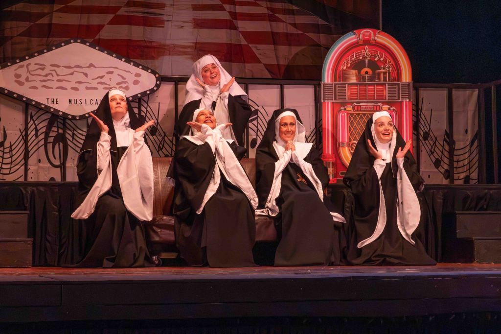 Five women dressed as nuns sitting on a stage, smiling and gesturing joyfully, with a jukebox and musical notes backdrop.