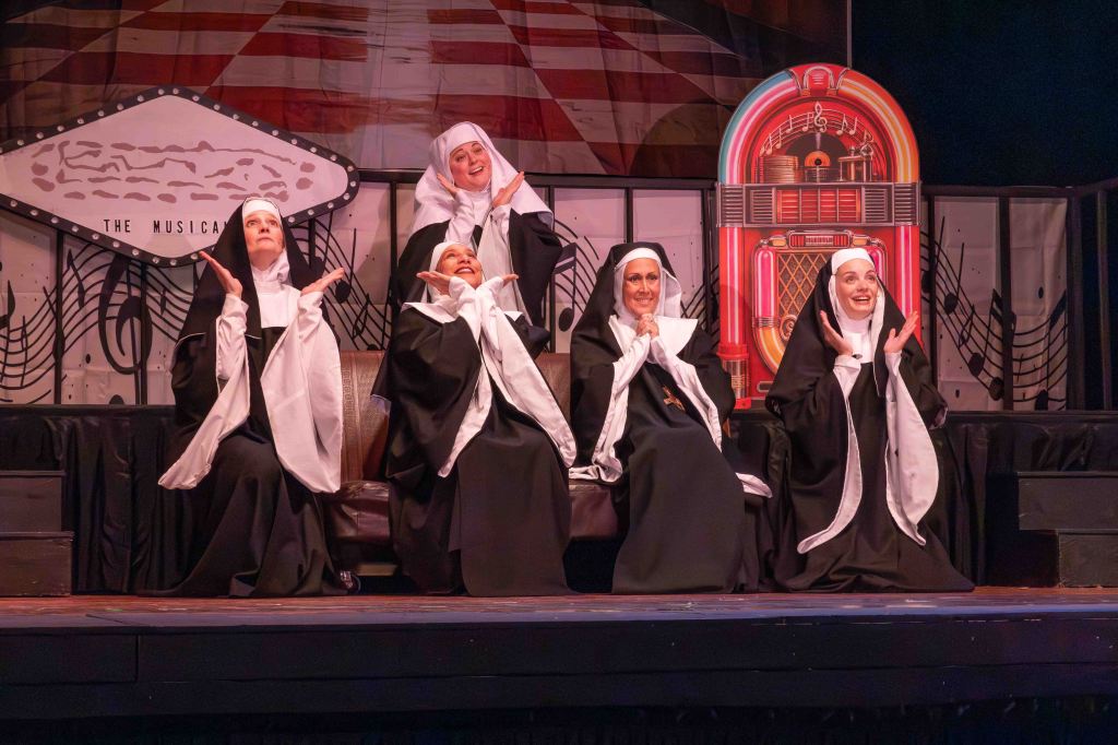 Five nuns in black and white costumes performing on stage with a colorful jukebox backdrop and musical notes.