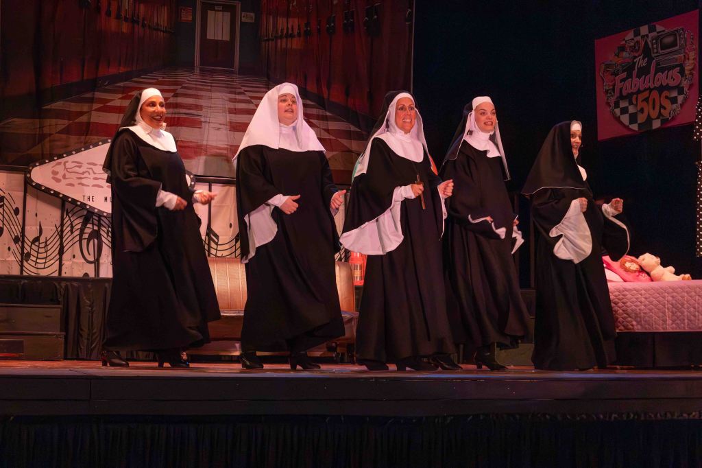 A group of five women in traditional nun costumes performing on stage. The background features a colorful 1950s theme with musical notes and a retro design.