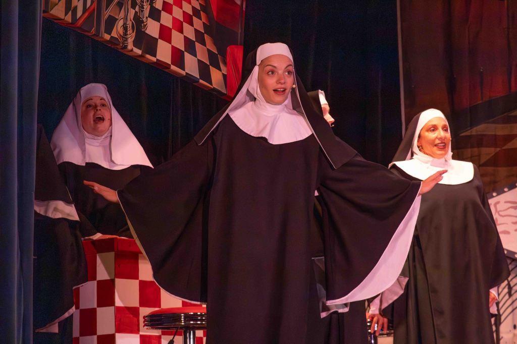 A group of nuns performing on stage, with one nun in the foreground gesturing dramatically while singing, and two other nuns visible in the background, creating a lively theatrical scene.
