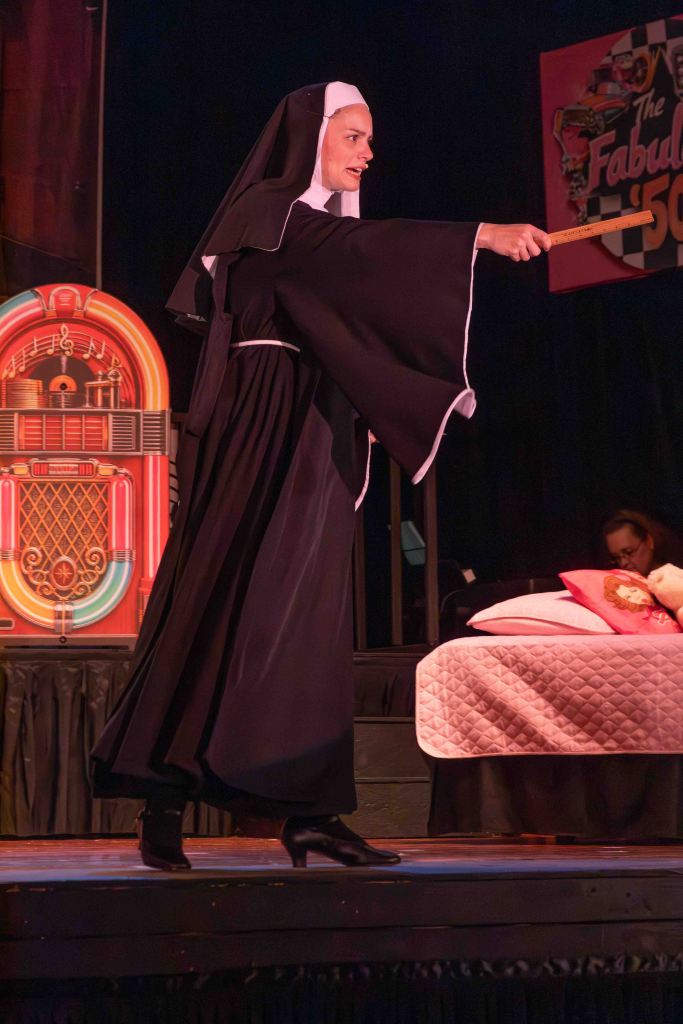 A performer dressed as a nun gestures dramatically on stage, wearing a traditional black habit and high heels, with a colorful jukebox and a bed with pillows in the background.