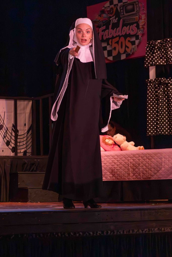 A performer dressed as a nun passionately delivering a speech on stage, with a pink bed and stuffed toys in the background, under a banner that reads 'The Fabulous '50s'.