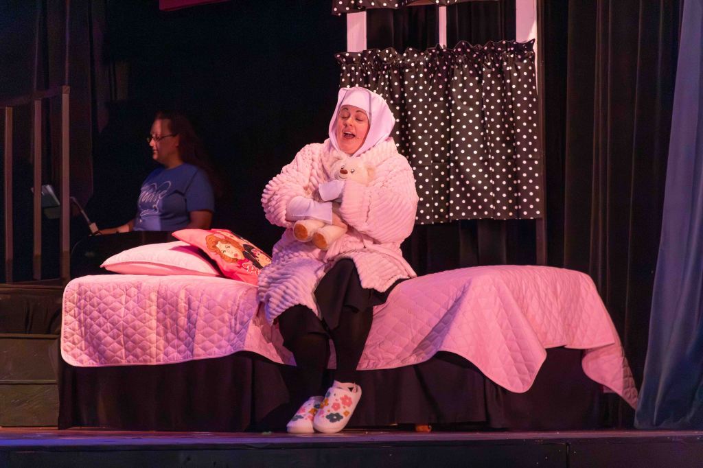 A woman in a cozy white robe and headscarf sits on a pink bed, holding a stuffed animal and singing. In the background, another woman is seen with a clipboard, participating in a stage performance.