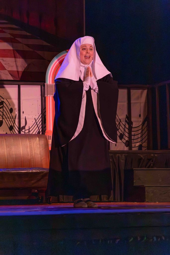 A performer dressed as a nun stands on stage, smiling and with hands clasped in prayer, in front of a jukebox backdrop.