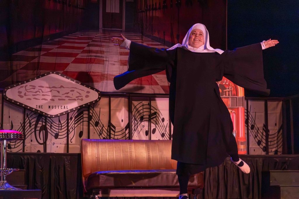 A performer dressed as a nun is dancing joyfully on stage, with arms outstretched, in front of a vibrant backdrop featuring musical notes and a jukebox.