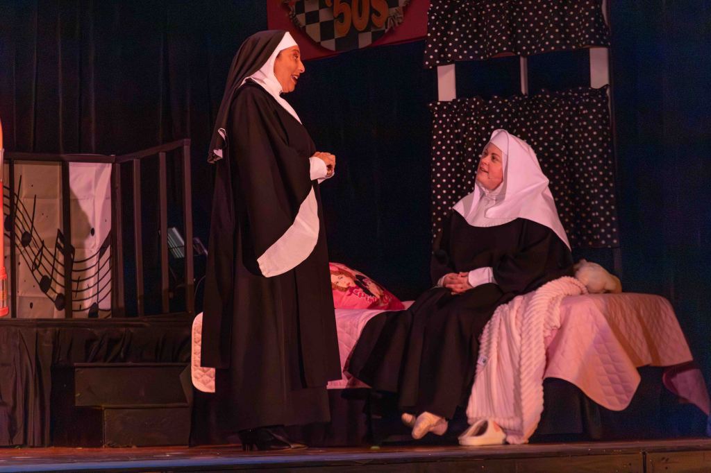 Two nuns in traditional attire on stage, one standing and speaking while the other sits on a bed with a blanket, set in a theatrical backdrop.