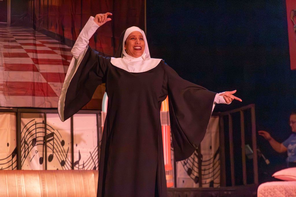 A woman dressed as a nun performs joyfully on stage, with her hand raised and a smile, in front of a colorful backdrop featuring musical notes.