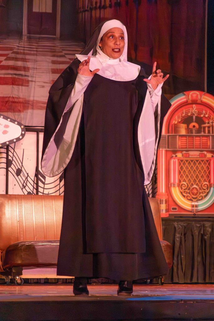 A woman dressed as a nun performs on stage, gesturing expressively in front of a vintage backdrop featuring musical notes and a jukebox.